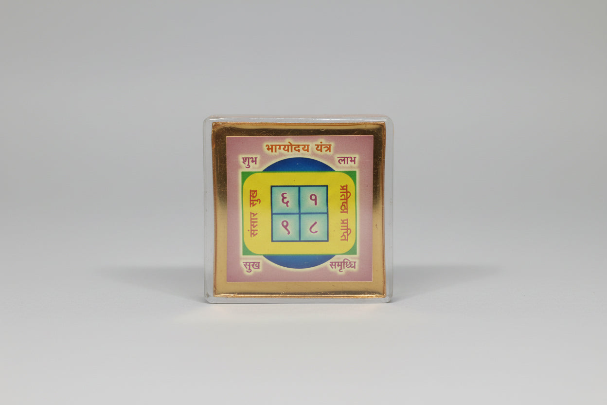 A colorful square-shaped Bhagyoday Yantra with numerical codes and sacred geometry symbols, predominantly pink and yellow with black text.