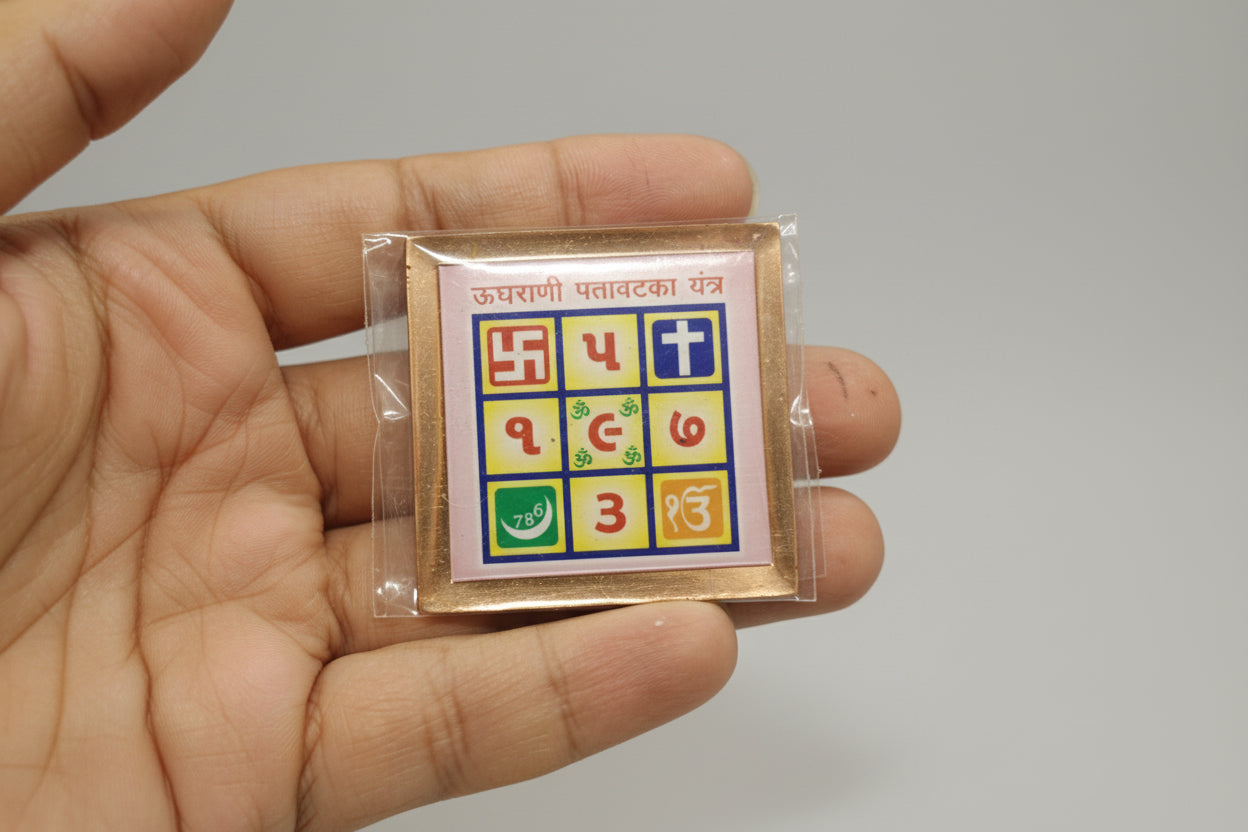 A hand holding a small yantra or spiritual symbol with geometric shapes and numbers, possibly used for recovering stuck money, against a plain background.