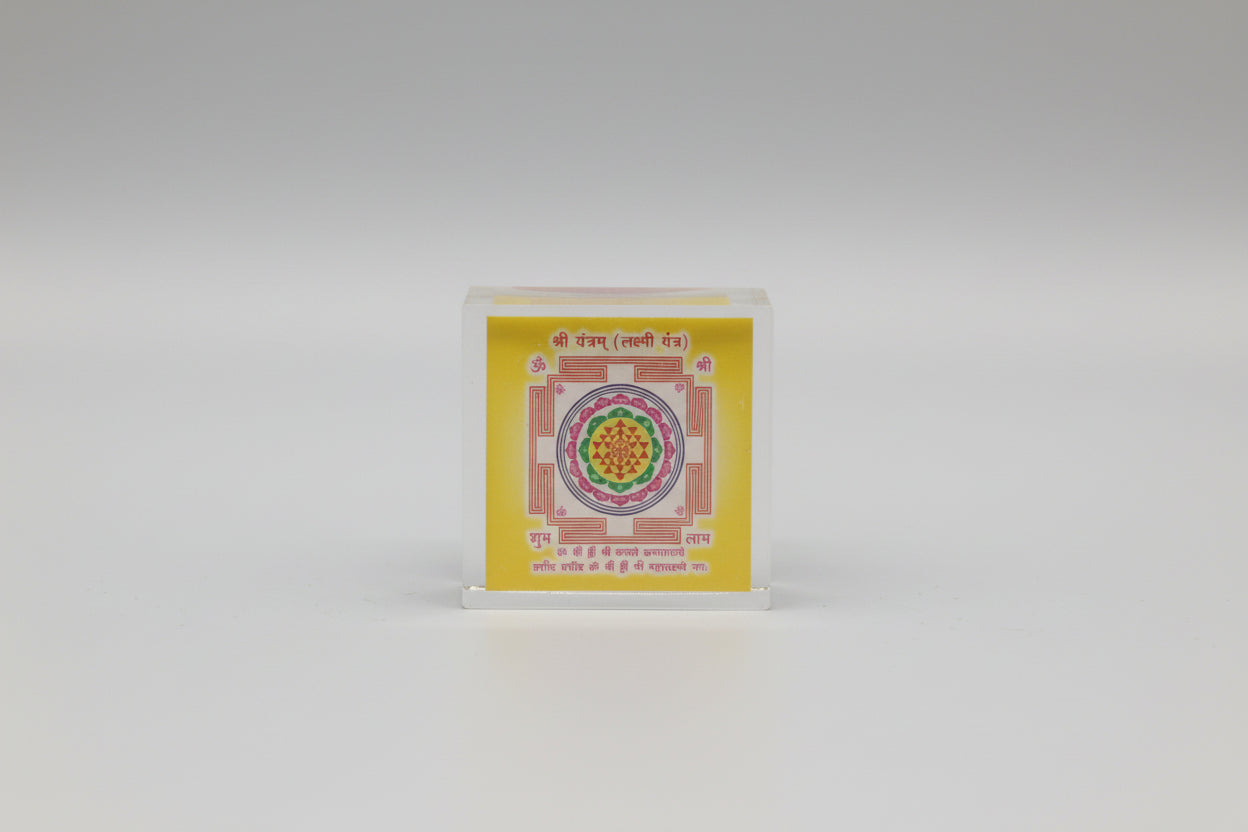 A hand holding a photo of a Shri Yantra (Laxmi Yantra) with a colorful geometric pattern on a yellow background, with Devanagari script at the top.