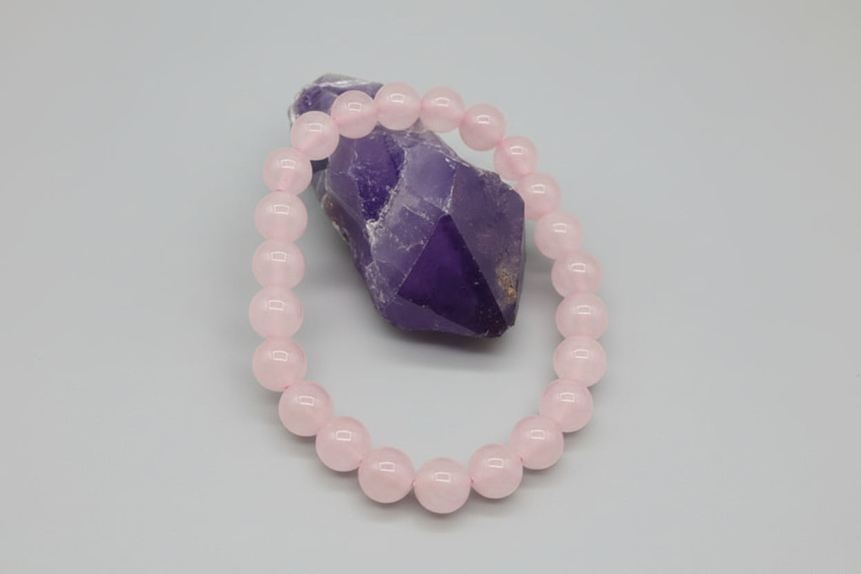 A heart-shaped rose quartz bracelet placed on a white surface with a larger rose quartz stone in the background.
