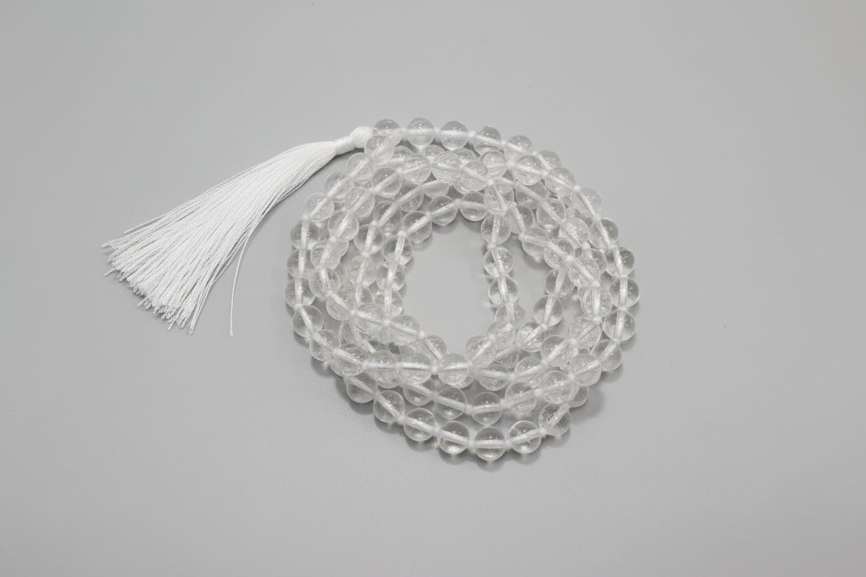 A clear quartz japmala with a red and white tassel, displayed on a red background. The japmala beads are translucent, and text on the image attributes its type and birthdate significance.