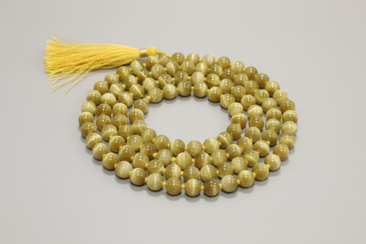 A Cat's Eye Japmala with green beads and a yellow tassel, laid out on a red surface. Text on the image indicates birthdate and Mahadasha information.