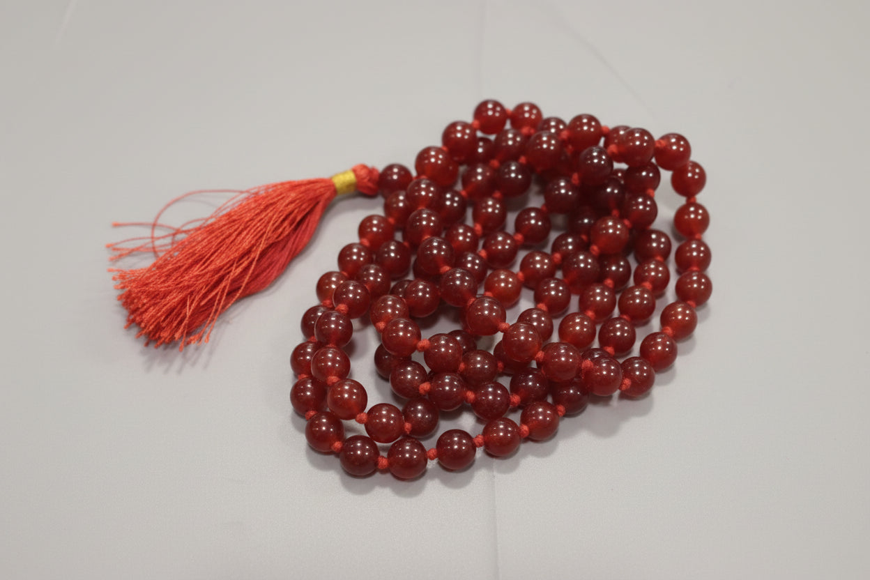 A Carnelian Japmala with red beads and a tassel, placed on a red surface, with the labels 'Carnelian japmala' and 'Strength and power' applied to it.
