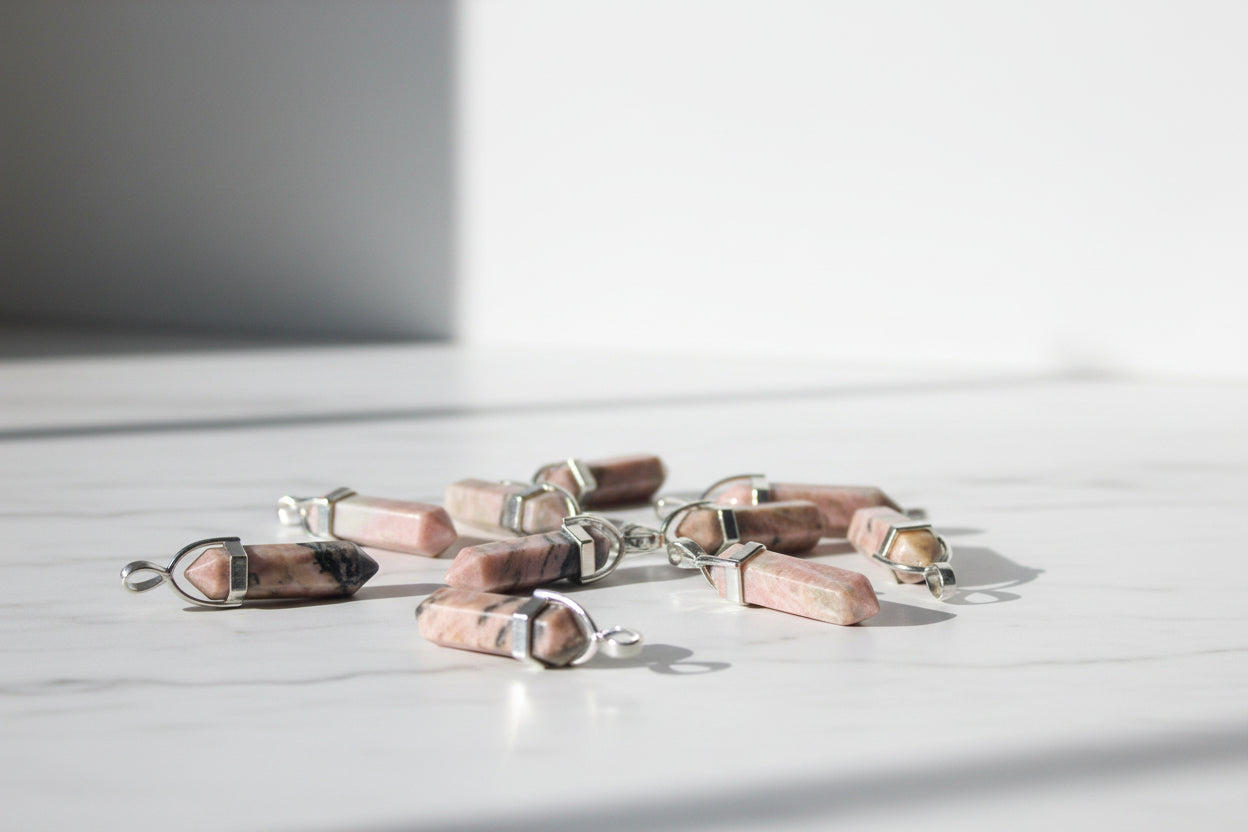 A collection of rhodonite pencil-shaped pendants with metal caps at the pointed ends, displayed on a textured brown surface.