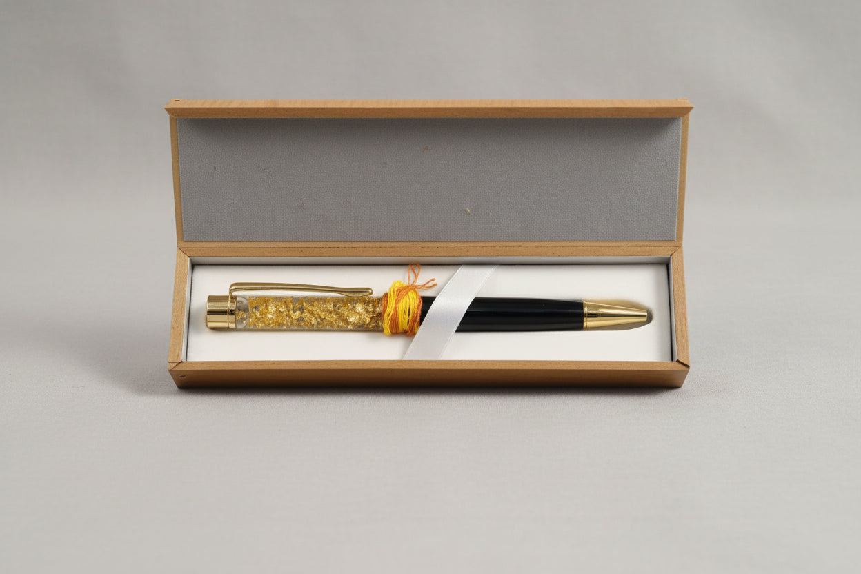 A Kanakdhara pen displayed in an open box, featuring a black body with a golden cap, a yellow string tied around it, and a golden decorative element in the nib section.