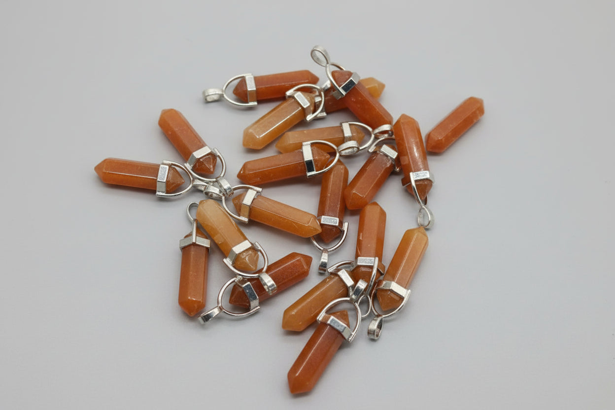 A collection of red aventurine pencil-shaped pendants with metal caps and loops, laid out on a textured surface.
