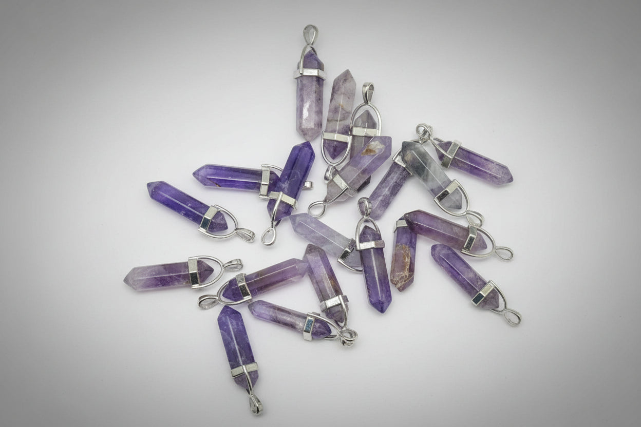 A collection of amethyst pencil-shaped pendants with metal caps at each end, displayed on a textured brown surface.