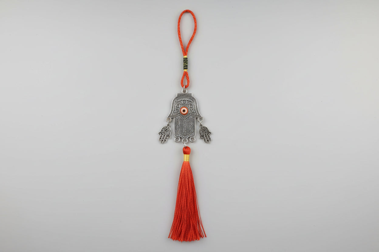 A metal Hamsa hand hanging with a red tassel, displayed against a textured background.